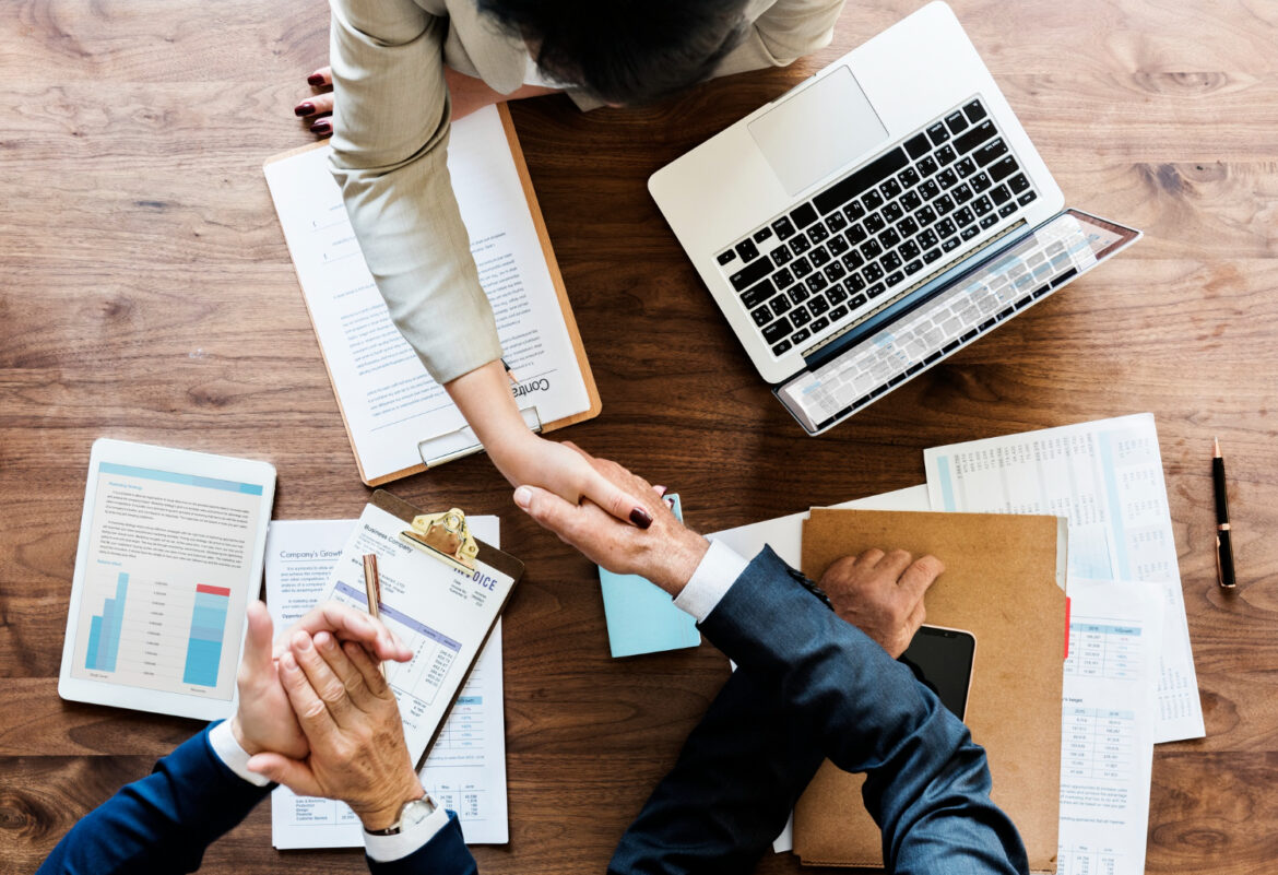 Top view of business professionals handshaking over a desk with laptop, tablet, and analytics charts, representing strategic AI integration in business.