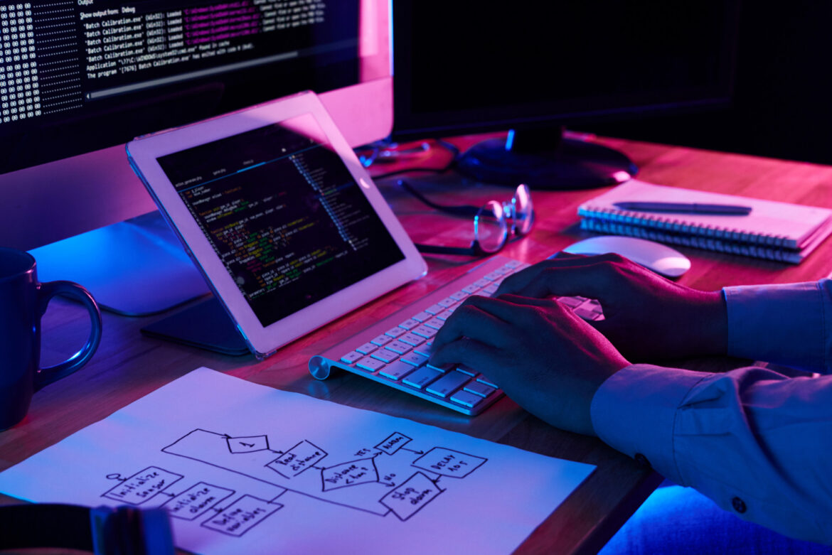 Developer coding custom web functionality on a keyboard with flowchart planning paper and dual monitors.